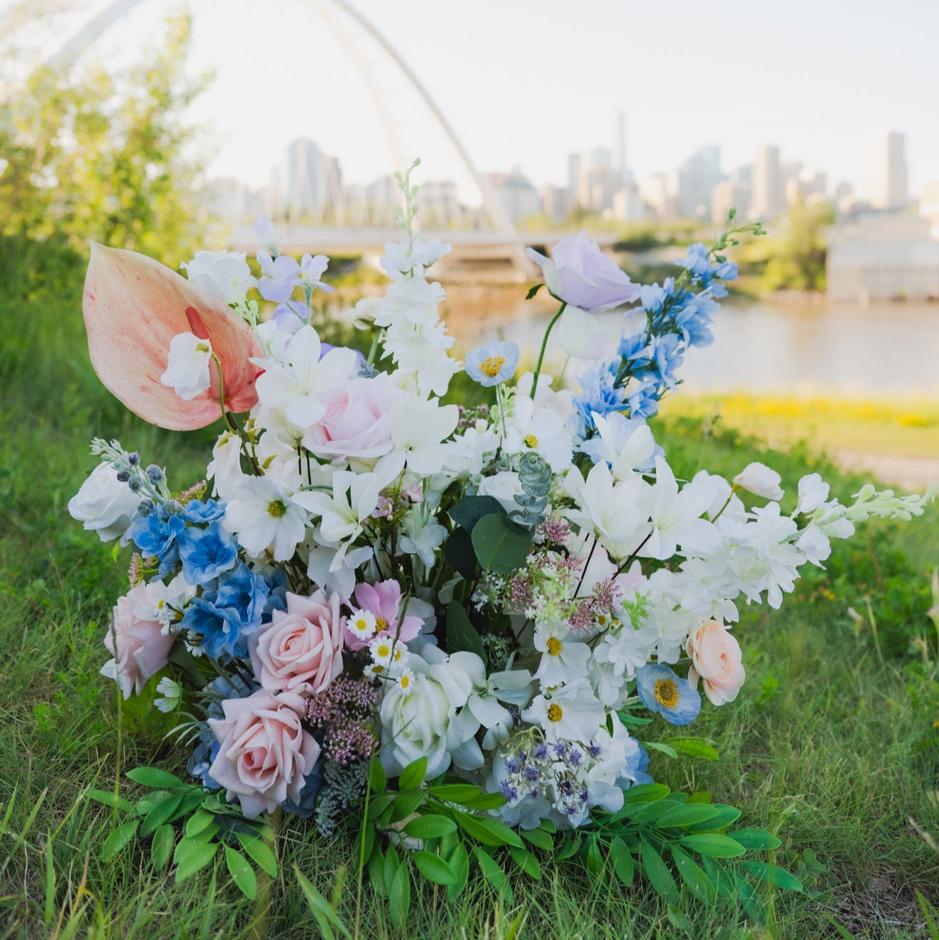 Artificial pastel floor floral arrangement with blush and lilac roses, white hydrangeas, and blue delphiniums placed near Edmonton’s Walterdale Bridge, perfect for wedding ceremony or reception rentals.