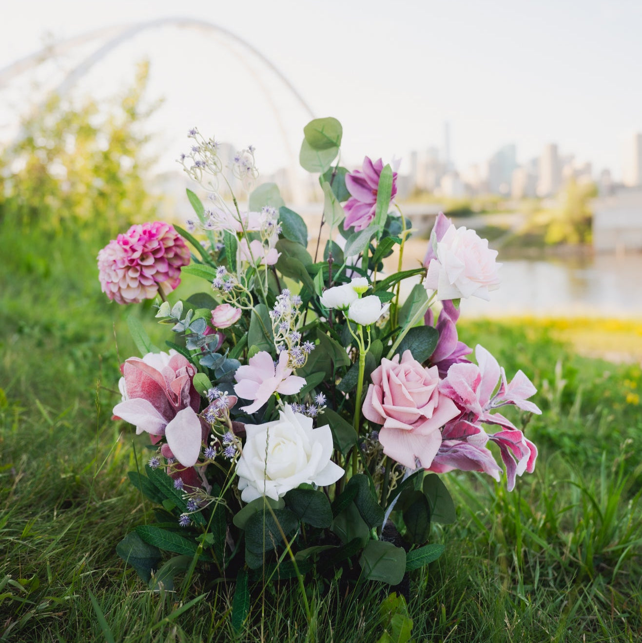 Plum Bliss Floor Florals. Aisle Markers. Floral arrangement in a basket on grass with a blurred cityscape in the background.