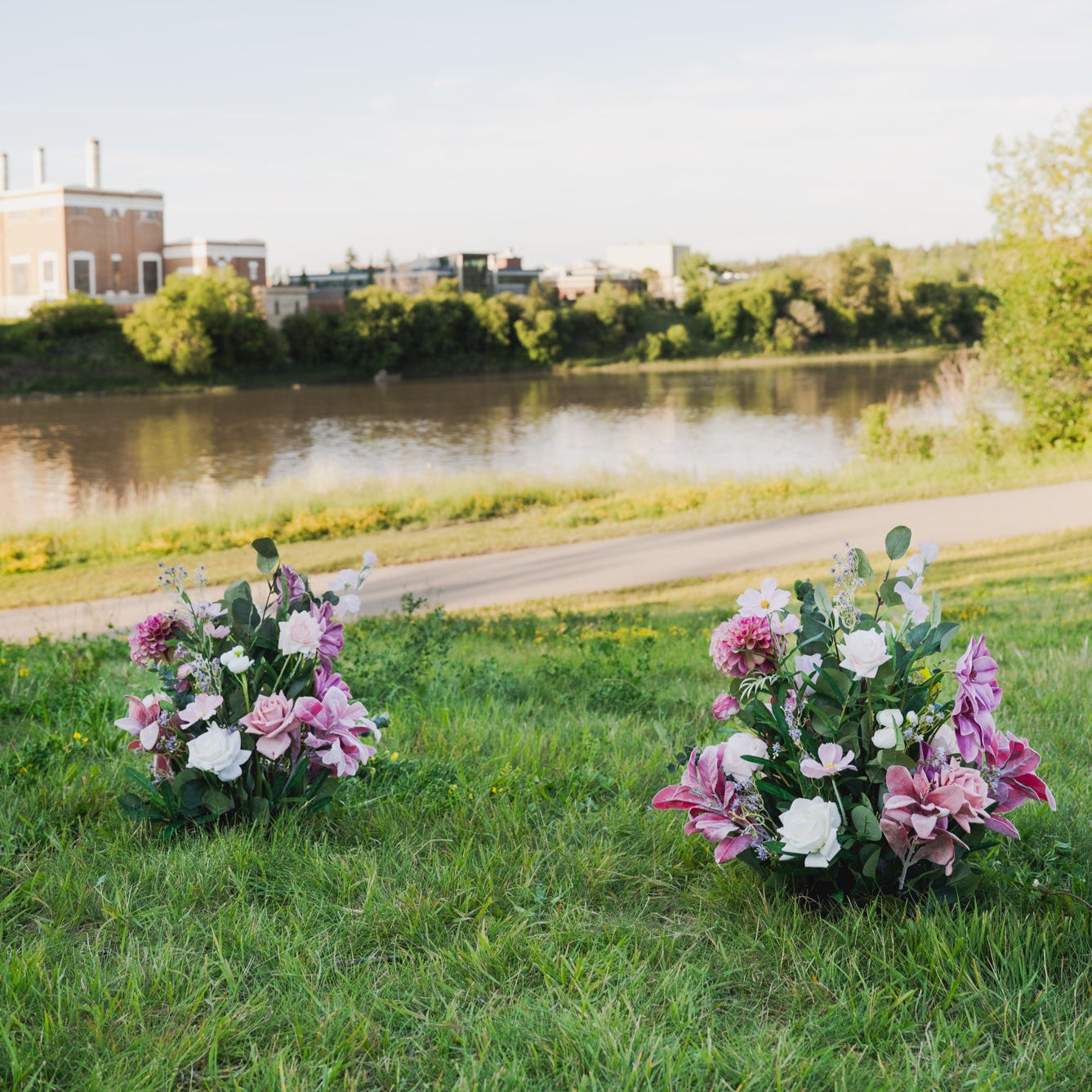 Two flower arrangements on a grassy area near a body of water with buildings in the background. Aisle markers of the Plum Bliss collection.
