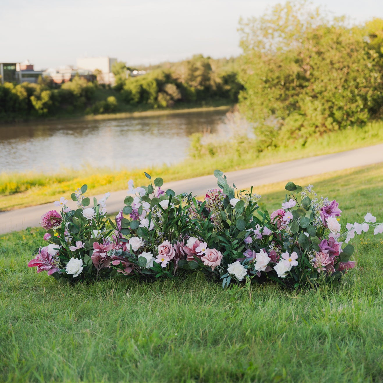 Floral arrangement on grass with a river and trees in the background. Plum Bliss Floor Florals (aisle markers) lined up on the grass.