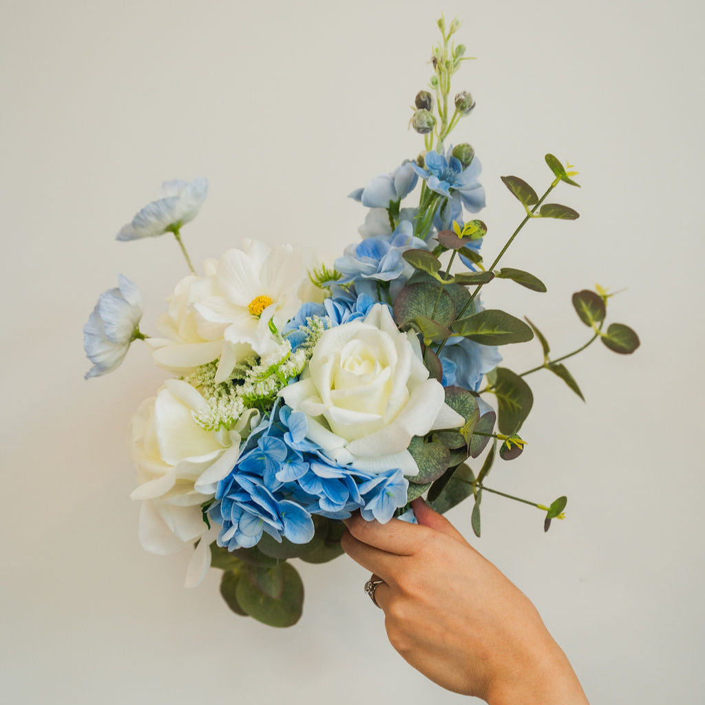 Hand holding the Ocean Breeze faux flower bridesmaids bouquet of blue and white flowers against a plain background for special events.