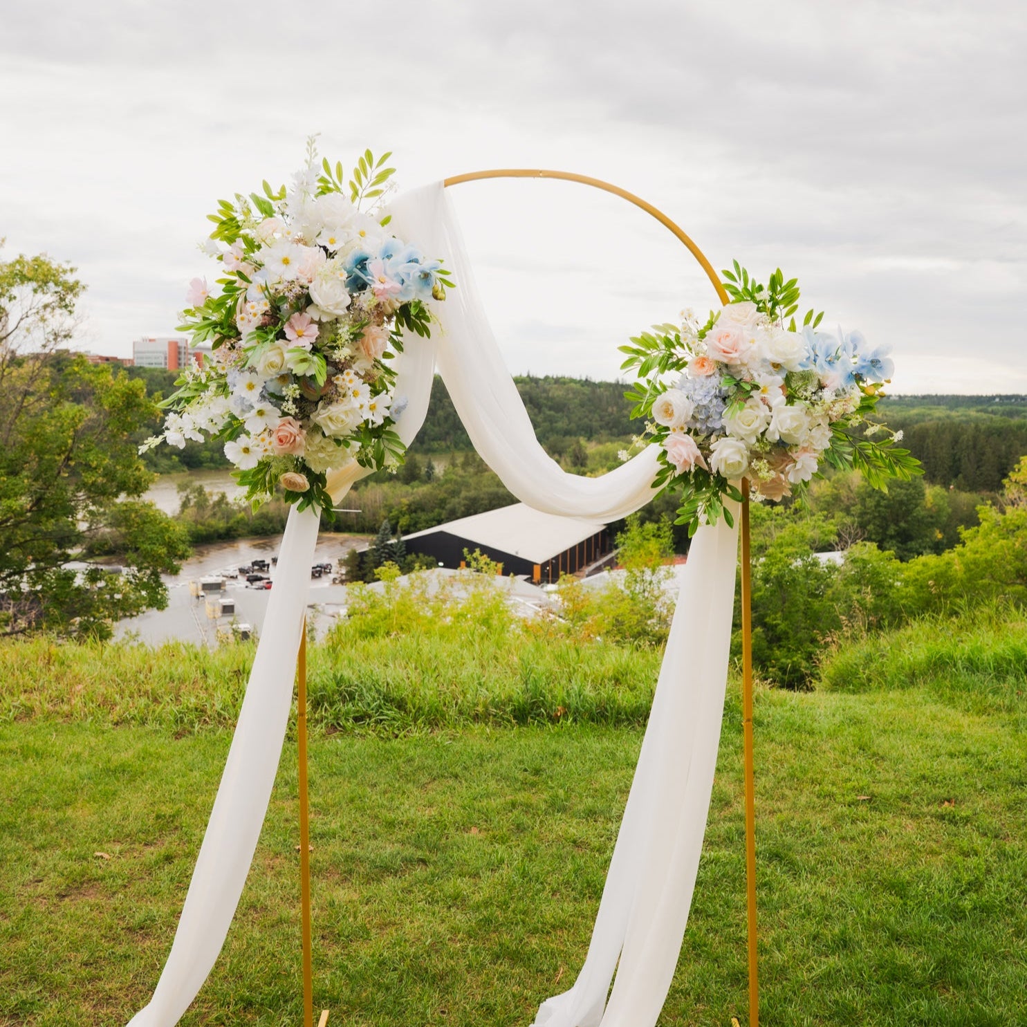 Faux wedding arch rental in Edmonton featuring artificial flowers in blush, ivory, and light blue with white draping on a gold arch stand, perfect for outdoor ceremonies.