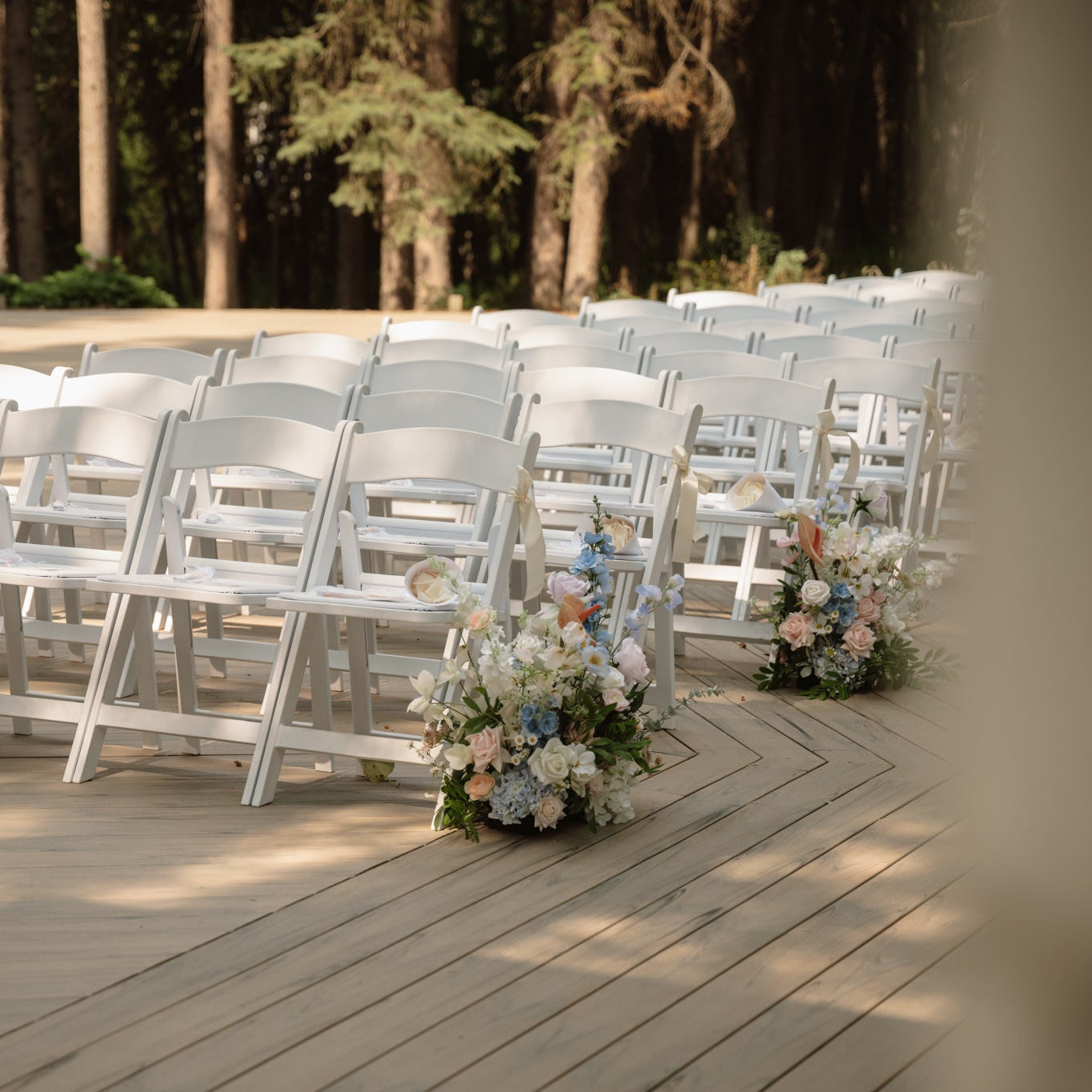 White chairs arranged on a wooden deck with floral arrangements, set against a forest backdrop.