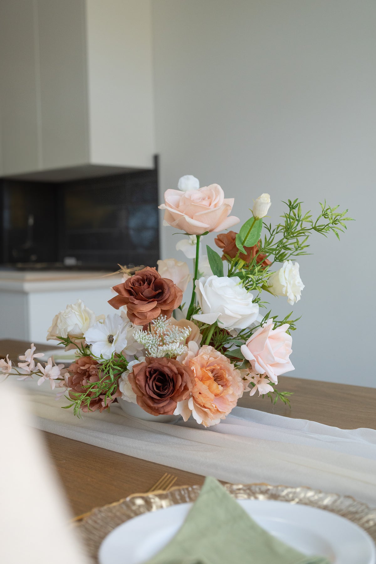 A rustic floral centerpiece with blush pink, terracotta, and white flowers arranged on a white tulle runner over a wooden table, with a blurred dining setting in the background.