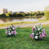 Two flower arrangements on a grassy area near a body of water with buildings in the background. Aisle markers of the Plum Bliss collection.
