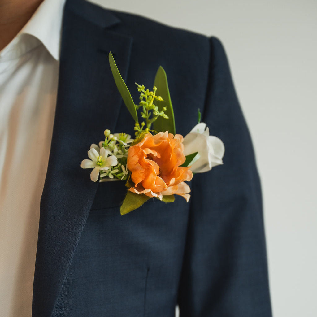 A close-up of the Cocoa Bloom Groomsmen’s Boutonniere on a navy suit jacket, featuring a soft cocoa-toned bloom with white accents and greenery.