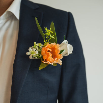 A close-up of the Cocoa Bloom Groomsmen’s Boutonniere on a navy suit jacket, featuring a soft cocoa-toned bloom with white accents and greenery.