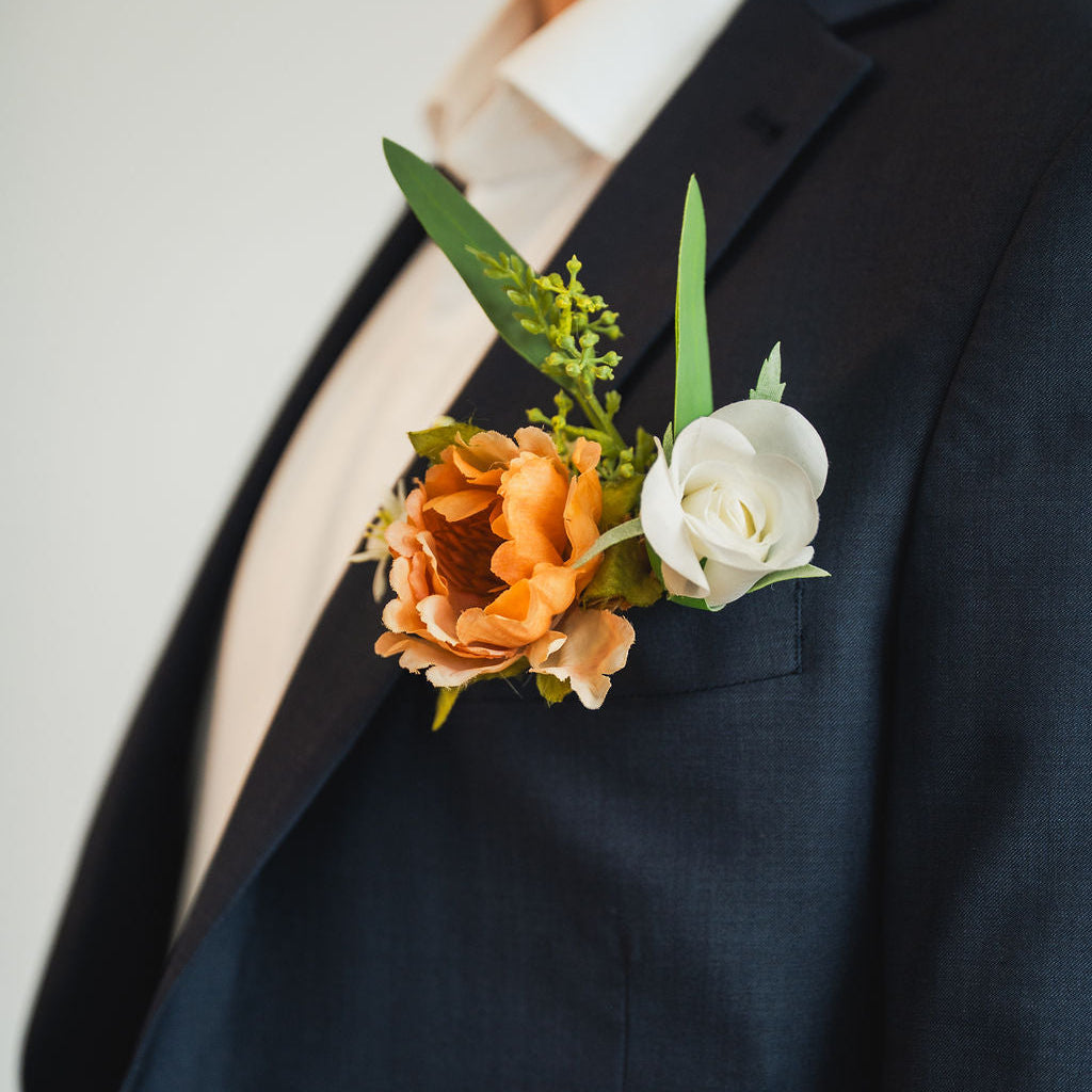 Side view of the Cocoa Bloom Groomsmen’s Boutonniere on a groomsmens navy suit, highlighting the warm cocoa flower, white rose, and leafy greens.