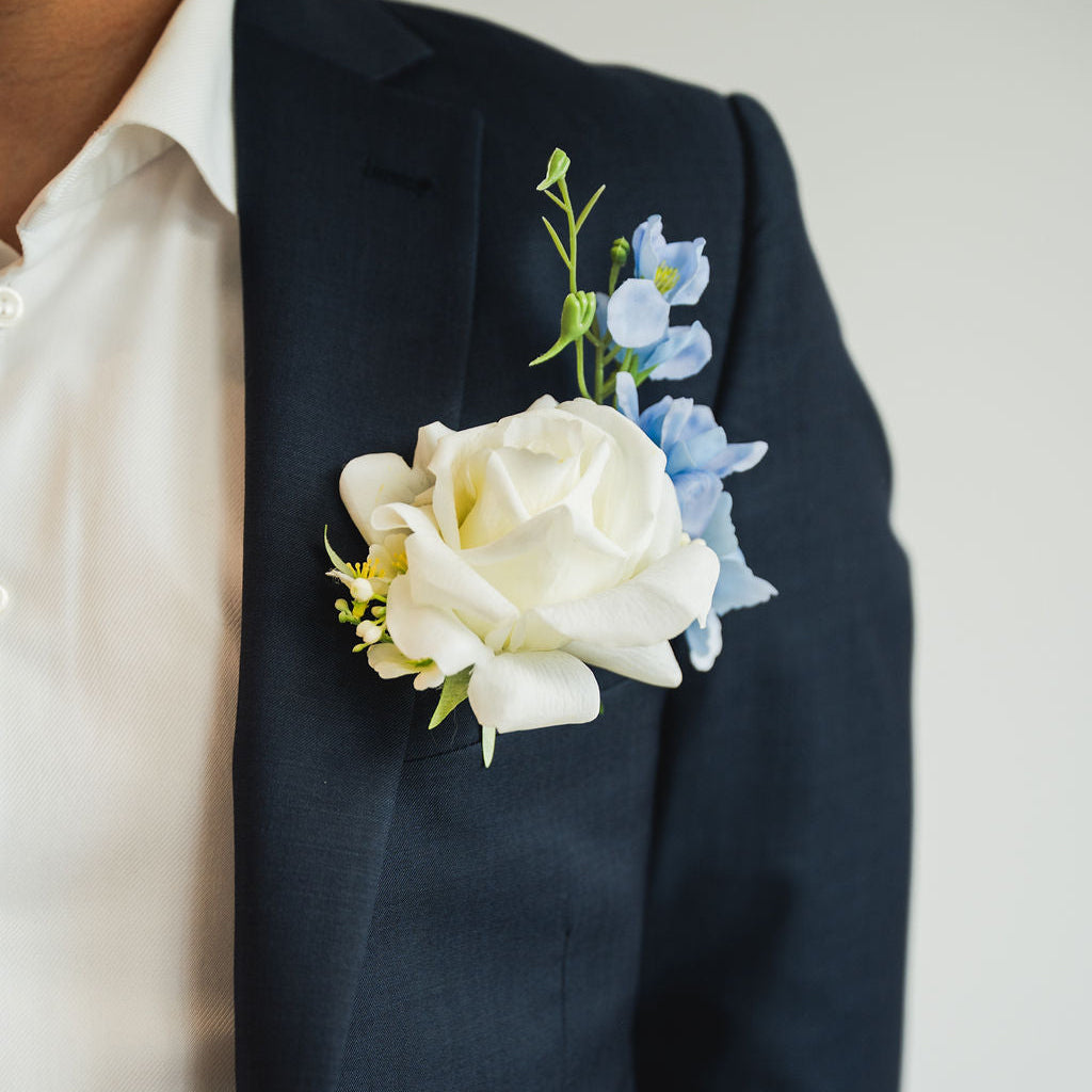 Navy blazer with an artificial, real tiuch white rose and blue boutonniere on a plain background for special events including weddings, grad and more.