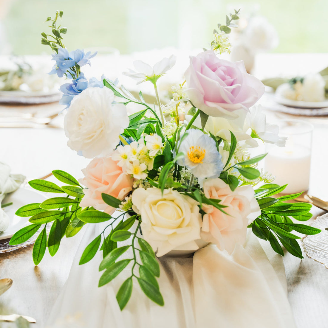 Artificial flower centerpiece with blush and ivory roses, blue faux blooms, and lush greenery, available for Edmonton wedding or special event rentals.