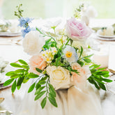 Artificial flower centerpiece with blush and ivory roses, blue faux blooms, and lush greenery, available for Edmonton wedding or special event rentals.