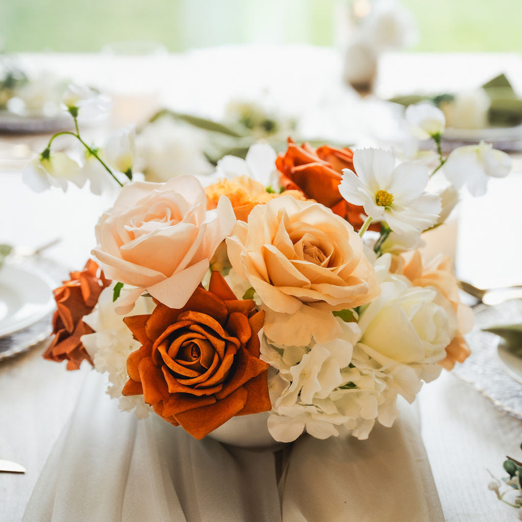 Silk and real-touch flower centrepiece with warm-toned roses in peach, caramel, and rust, accented by ivory blooms, displayed on a dining table setting.