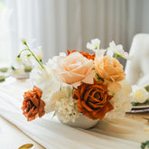 Artificial floral centrepiece featuring caramel, ivory, and copper-toned faux roses arranged in a white vase, styled on a wooden table with a soft ivory table runner.