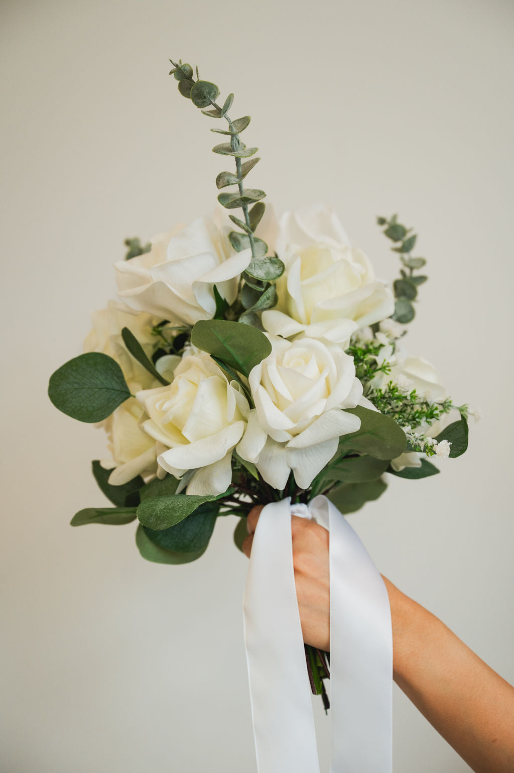 Bouquet of an artificial white bridal bouqet with green leaves held by a hand against a plain background