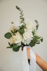 Bouquet of an artificial white bridal bouqet with green leaves held by a hand against a plain background