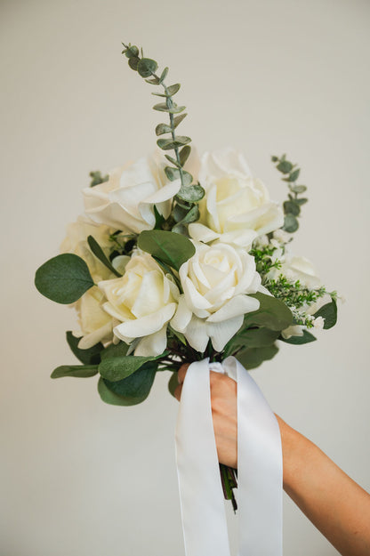 Bouquet of an artificial white bridal bouqet with green leaves held by a hand against a plain background
