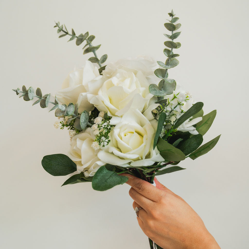 Bouquet of faux white roses and greenery held by a hand against a plain background