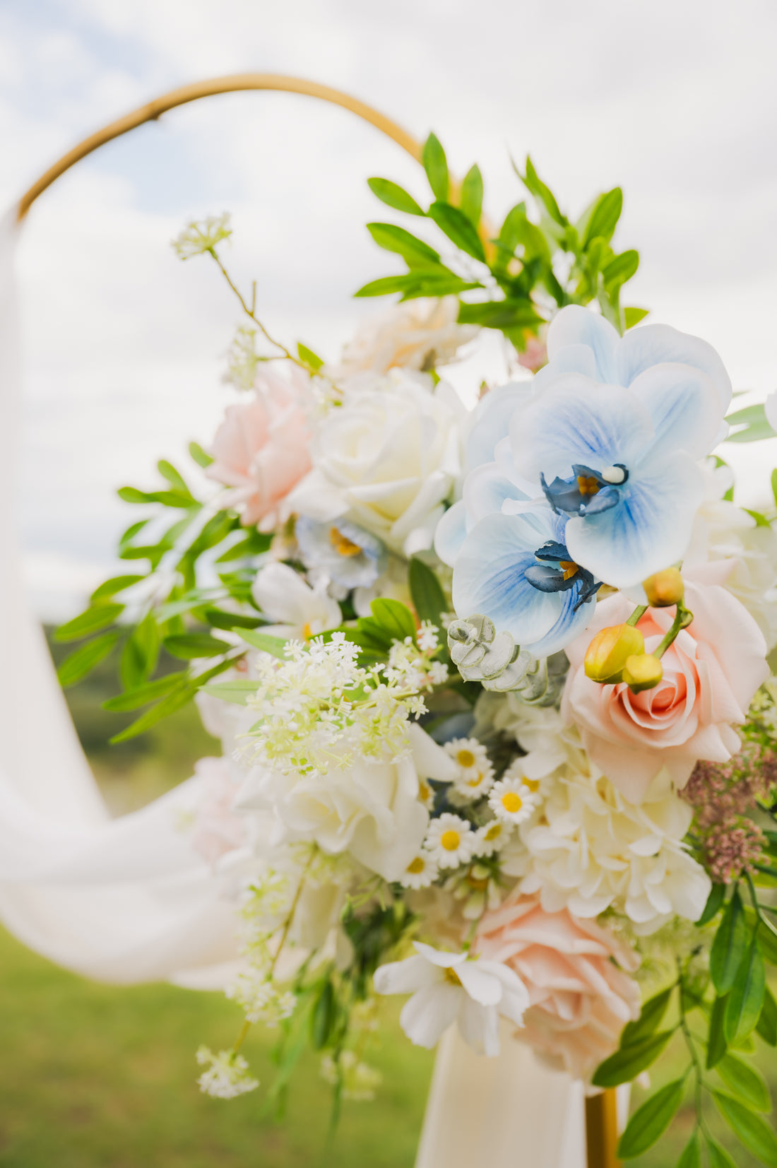 Side view of pastel wedding arch arrangement with artificial blue orchids, blush roses, ivory flowers, and greenery cascading from a gold circular arch.