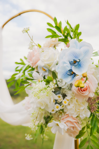 Side view of pastel wedding arch arrangement with artificial blue orchids, blush roses, ivory flowers, and greenery cascading from a gold circular arch.