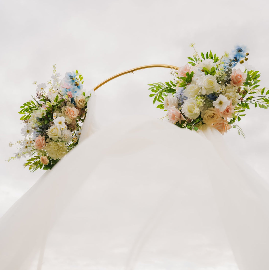 Wedding arch with white draping and pastel faux floral swags in ivory, blush, and blue tones on a gold circular stand, viewed from below against the sky.