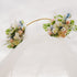 Wedding arch with white draping and pastel faux floral swags in ivory, blush, and blue tones on a gold circular stand, viewed from below against the sky.