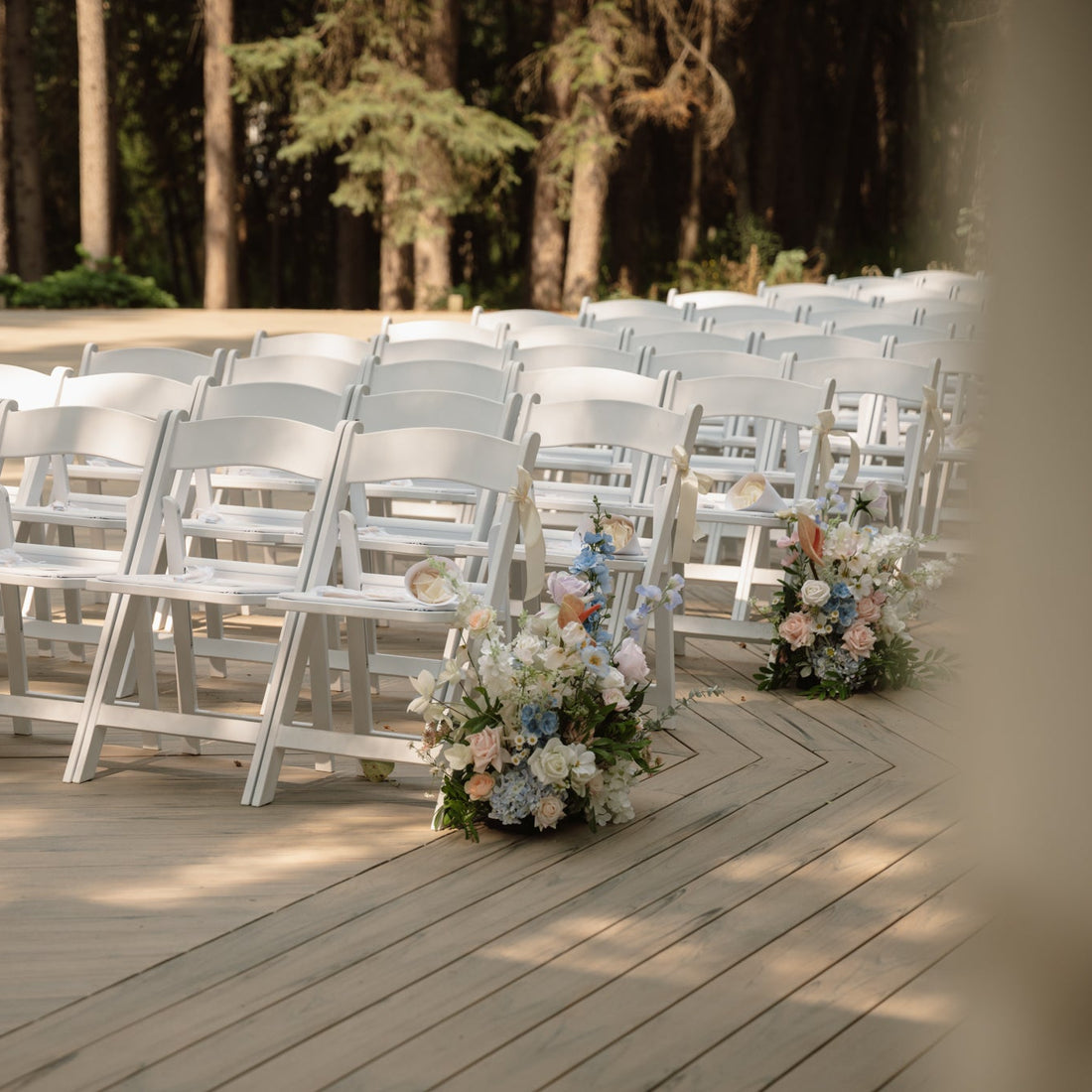 White chairs arranged on a wooden deck with floral arrangements, set against a forest backdrop.