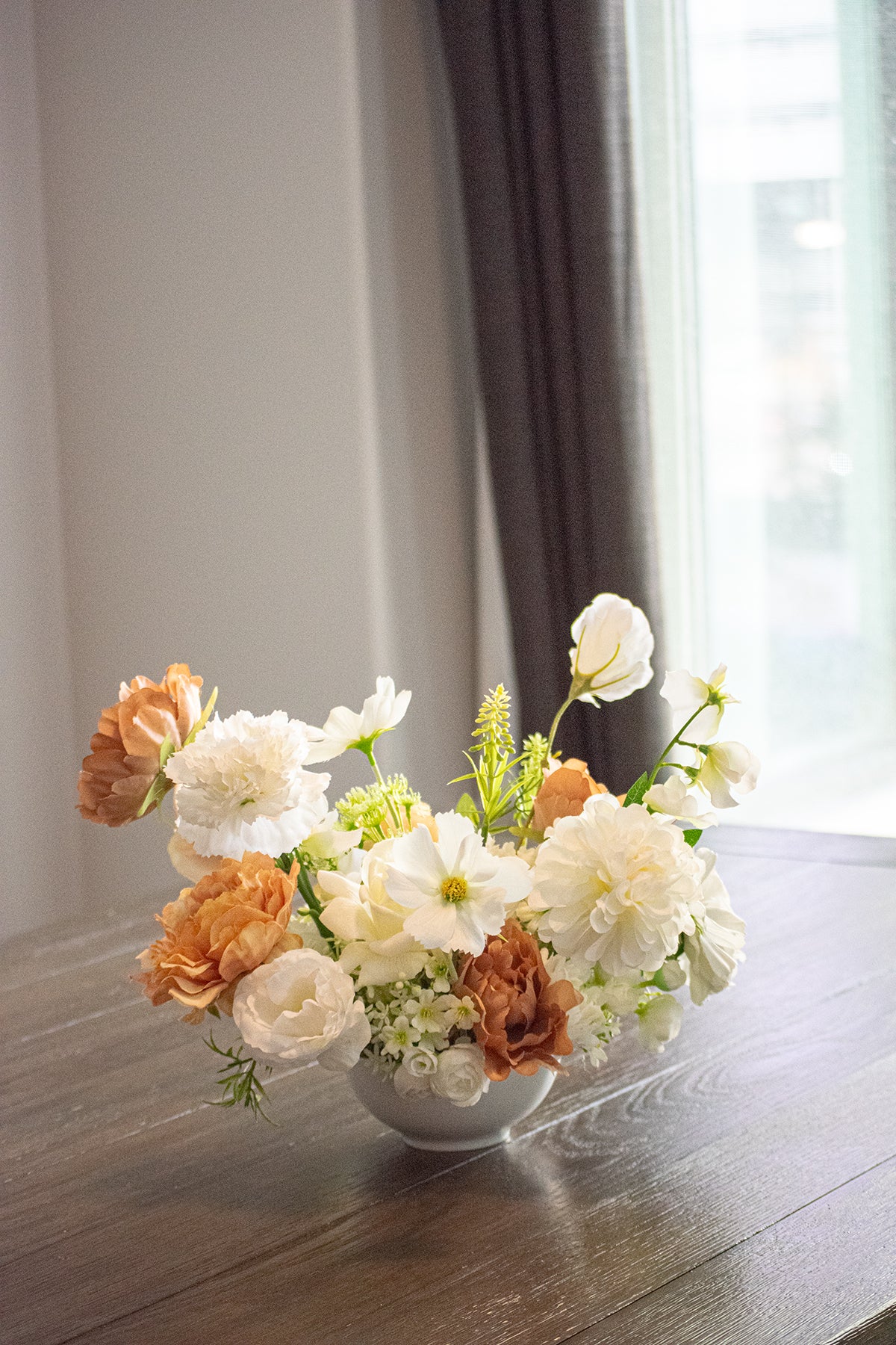 Floral arrangement in a white vase on a wooden table with a blurred window and curtain in the background.