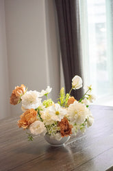 Floral arrangement in a white vase on a wooden table with a blurred window and curtain in the background.