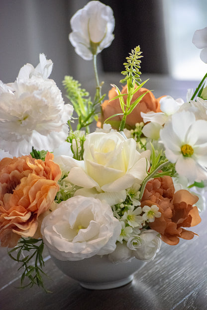 Close up of a faux flower table centrepiece with white, brown and orange flowers in a white vase on a wooden surface.