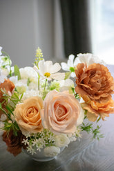 Close up of light brown, peach, blush and white faux flowers (silk and real touch) in a white vase on a reflective surface.