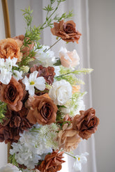 A close-up of a decorative floral arrangement featuring faux roses and other flowers in warm brown, peach, white, and beige tones against a soft light gray background.