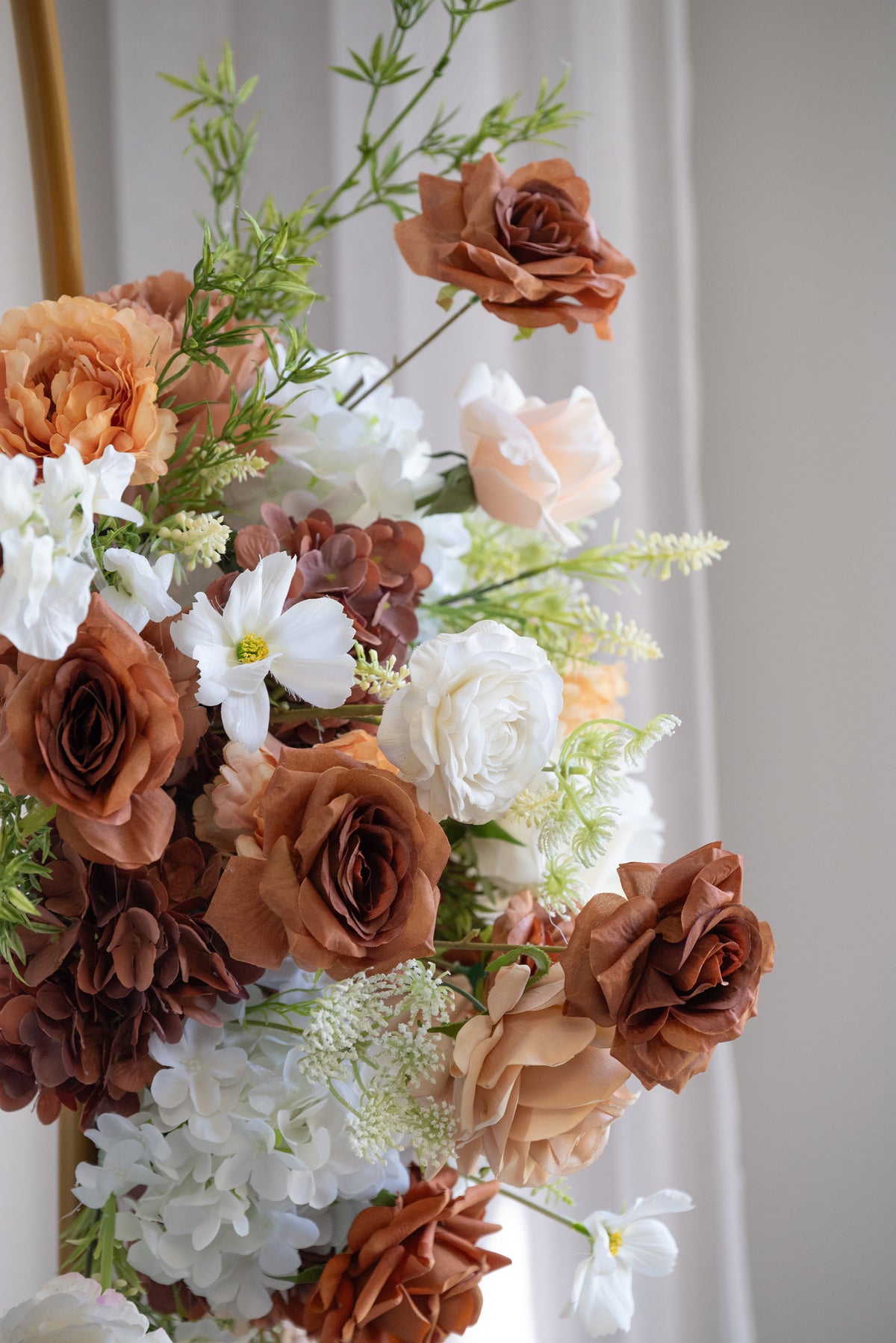 A close-up of a decorative floral arrangement featuring faux roses and other flowers in warm brown, peach, white, and beige tones against a soft light gray background.