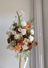A large artificial floral arrangement in earthy tones of terracotta, blush, cream, and white, displayed on a wooden stand against a neutral light gray wall and curtain background.