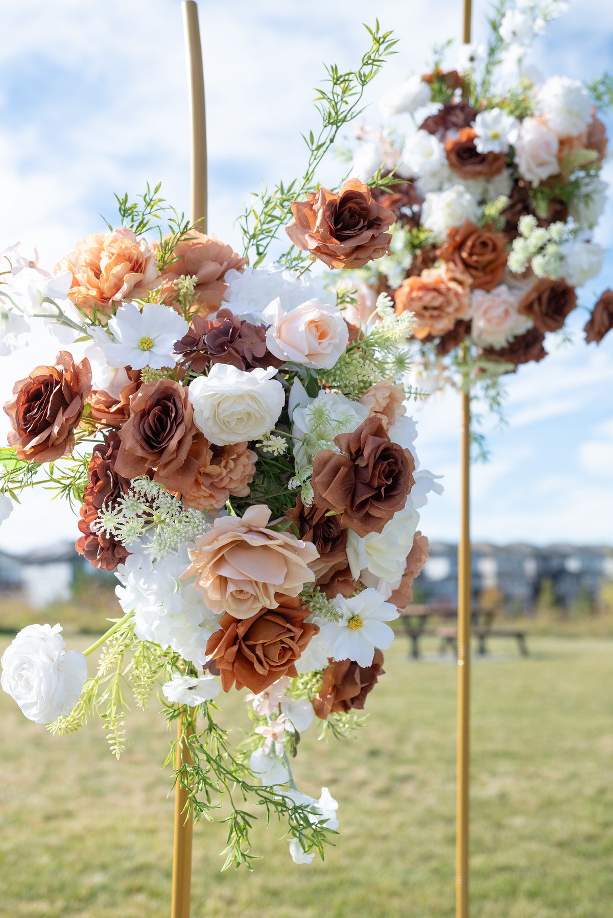 A large floral arch decoration with white, peach, and brown faux roses, green foliage, and gold metal poles set outdoors on a grassy field under a blue sky.