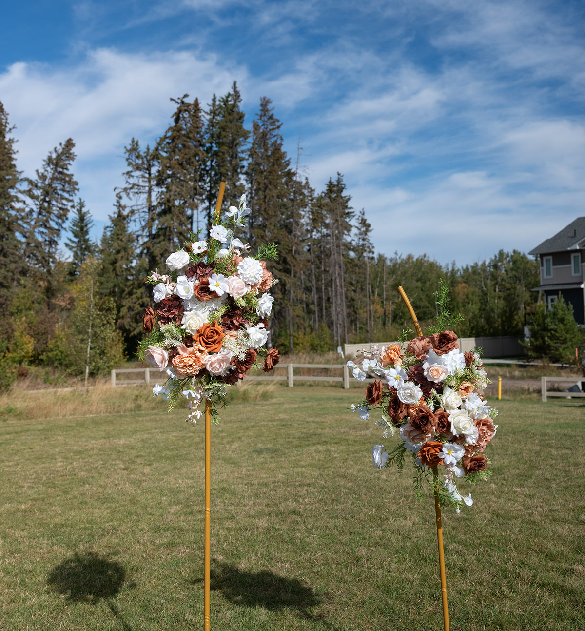 Two rustic floral arrangements on wooden poles set in a grassy outdoor field, with trees and a house in the background under a blue sky.