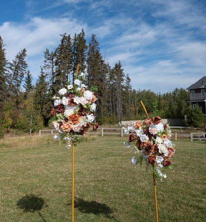 Two rustic floral arrangements on wooden poles set in a grassy outdoor field, with trees and a house in the background under a blue sky.