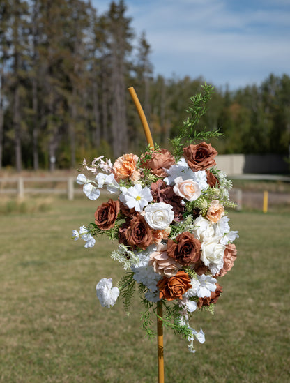 A rustic floral arrangement featuring white daisies, brown and peach roses, and green foliage, mounted on a wooden stake outdoors in a grassy field with trees in the background.