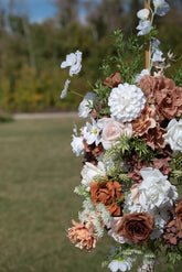 A close-up of a decorative floral arrangement featuring white, blush pink, and terracotta faux flowers with green foliage, set against a blurred outdoor grassy background.