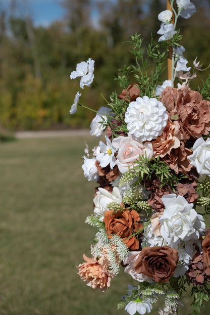 A close-up of a decorative floral arrangement featuring white, blush pink, and terracotta faux flowers with green foliage, set against a blurred outdoor grassy background.