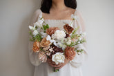 A person wearing a white off-the-shoulder dress holds a large rustic-style floral bouquet with white, peach, tan, and brown flowers against a plain light gray wall.