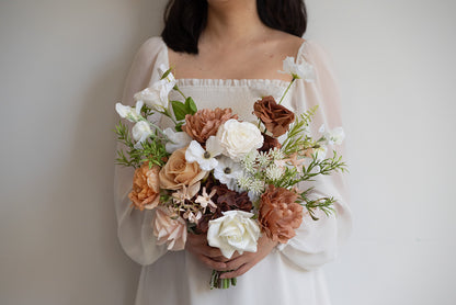A person wearing a white off-the-shoulder dress holds a large rustic-style floral bouquet with white, peach, tan, and brown flowers against a plain light gray wall.