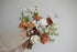 A hand holding a rustic-style floral bouquet with peach, white, and brown flowers and green foliage against a plain light gray background.