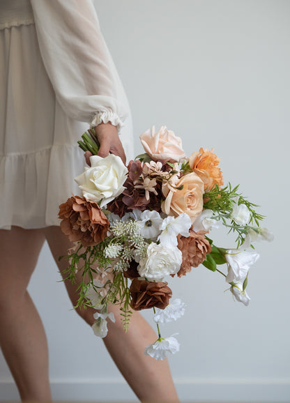 A person in a cream-colored dress holds a rustic, earthy-toned faux flower bouquet with white, peach, and brown blooms, set against a plain light gray wall.