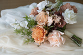 A bridal bouquet of artificial flowers and greenery, featuring blush pink, peach, and white blooms with white fabric draping underneath.