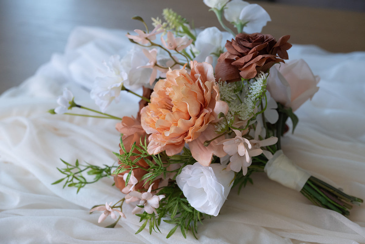 A rustic-style bridal bouquet featuring peach peonies, white ranunculus, a dark brown rose, and green foliage, resting on draped white tulle fabric.
