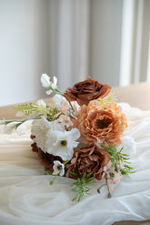 A rustic dried flower bouquet featuring brown roses, peach peonies, white anemones, and green foliage, arranged on draped white fabric.