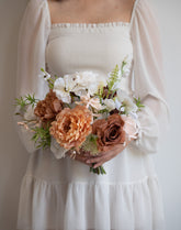 A person wearing a white smocked dress holds a bouquet of peach, white, and brown flowers with green foliage against a plain light background.