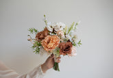 A hand holding a rustic-style floral bouquet with peach peonies, mauve-brown roses, white blossoms, and green foliage against a plain light gray background.