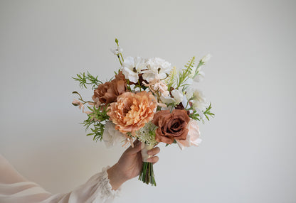 A hand holding a rustic-style floral bouquet with peach peonies, mauve-brown roses, white blossoms, and green foliage against a plain light gray background.