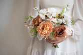 A close-up of a person in a white dress holding a rustic-style floral bouquet with peach, white, and brown flowers and green foliage.
